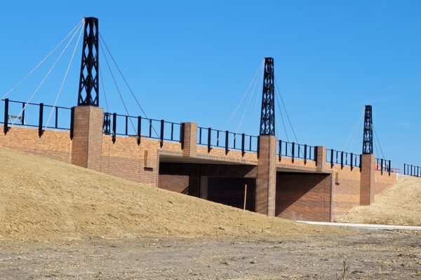 A brown bridge over a roadway with a blue sky. The bridge has 3 black structural columns and guardrails.