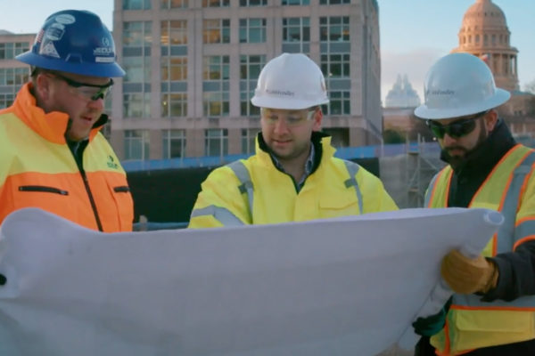 3 CobbFendley employees looking at a plan set in front of a building. All wearing hard hats and yellow safety vests.